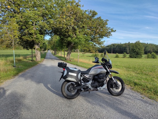 A Moto Guzzi V85 TT Travel 2022 parked on a road, at the far end of a tree lined road 